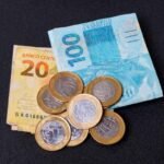 Cédulas de R$ 100 e R$ 20 acompanhadas de moedas de real sobre superfície escura, representando recebimento de valores, pagamento de precatórios, RPVs, créditos judiciais e recursos financeiros.