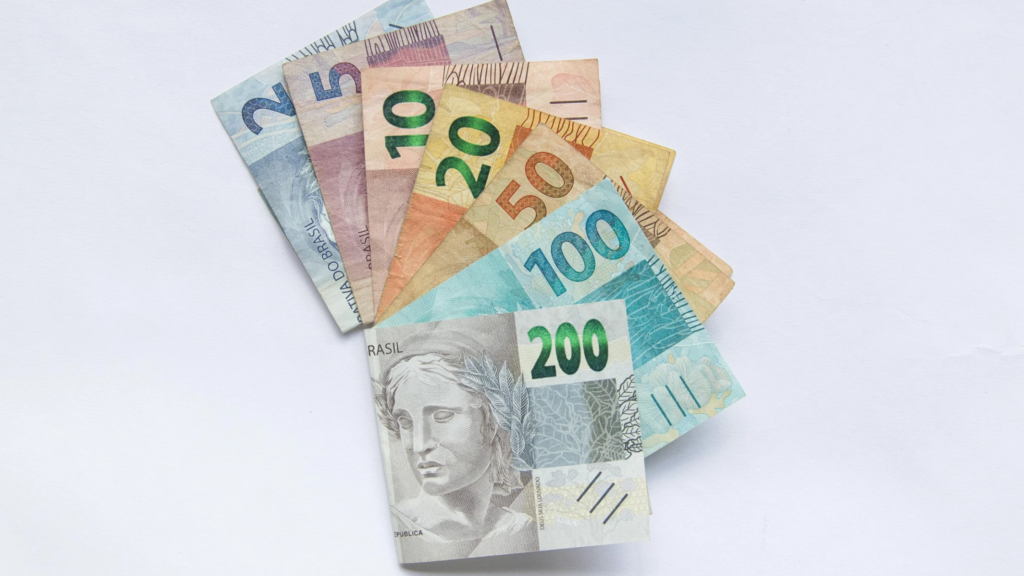 Conjunto de cédulas de real brasileiro disposto em leque sobre uma superfície branca. As notas visíveis são de R$ 2, R$ 5, R$ 10, R$ 20, R$ 50, R$ 100 e R$ 200, parcialmente sobrepostas e organizadas de forma a destacar seus valores. A imagem tem iluminação suave e fundo limpo, transmitindo conceitos relacionados a dinheiro, finanças, investimentos, pagamentos, economia e crédito.
