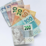 Conjunto de cédulas de real brasileiro disposto em leque sobre uma superfície branca. As notas visíveis são de R$ 2, R$ 5, R$ 10, R$ 20, R$ 50, R$ 100 e R$ 200, parcialmente sobrepostas e organizadas de forma a destacar seus valores. A imagem tem iluminação suave e fundo limpo, transmitindo conceitos relacionados a dinheiro, finanças, investimentos, pagamentos, economia e crédito.